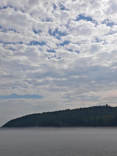 The image depicts a serene natural landscape. The foreground features a body of water with subtle ripples, possibly a river or lake. A thick layer of fog hovers just above the water's surface, obscuring the lower portion of the distant shore. The middle ground is dominated by a densely forested hillside, which rises steeply from the fog-shrouded water. The trees are a deep green, and the outline of the treeline forms a distinct curve against the sky. The sky above is filled with a complex pattern of white and gray clouds, with patches of blue visible in between. The overall impression is one of tranquility and natural beauty. The location is the city of Atamanovo, Russia.