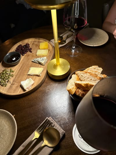 The image captures an intimate, dimly lit dining scene, likely in a restaurant or wine bar, centered around a cheese and bread platter accompanied by red wine. The setting is dark and warm, with artificial lighting creating highlights on various elements.

In the foreground and center, an oval wooden serving board presents an assortment of cheeses, fruits, seeds, and a dip. There are at least five different types of cheese, varying in color from pale yellow to off-white, and in texture, including a hard yellow cheese, a blue cheese with visible marbling, and softer white cheeses. Clustered on one side of the board are dark dried cranberries or cherries, and on the opposite side, a scattering of green pumpkin seeds. A small, dark gray bowl contains a dark, jam-like substance.

To the upper right of the cheese board, a small, dark bowl holds several slices of rustic-looking bread, likely a baguette. A wine glass filled with red wine is partially visible in the upper right foreground, obscuring part of the bread bowl. Another wine glass, also containing red wine, is positioned to the left of the cheese board.

A prominent feature in the center of the image is a modern, minimalist table lamp with a brass or gold-finished stem and base, emitting a warm, soft light that illuminates the items on the cheese board. The lamp's stem has a textured section.

The table surface is a dark brown, possibly polished wood, reflecting some of the light. To the far right, a set of gold-colored cutlery, consisting of a spoon and a fork, rests neatly on a light brown or beige linen napkin. Below the cutlery, a light-colored, textured ceramic bowl is partially visible.

A white circular coaster is visible to the right of the bread bowl and behind the foreground wine glass. The word "VIENTIANE" is clearly printed on it, along with a partial letter "S" above it and a stylized logo. Another circular coaster, darker and possibly cork, is visible in the top left background. A person's arm and hand are faintly visible in the upper left corner of the image, suggesting companionship.

The overall atmosphere is one of relaxed enjoyment, characteristic of an evening meal or social gathering.