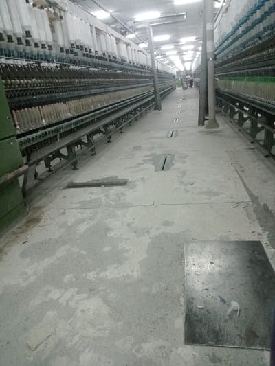 The image captures the interior of a textile or yarn spinning factory located in Sukomoro, Indonesia. A long, well-lit concrete aisle extends from the foreground into the deep background. This aisle is bordered on both sides by extensive rows of industrial spinning machinery. The machines on the left side are constructed with green metal frames, supporting numerous tiers of white yarn bobbins. Similarly, the machines on the right side display bobbins predominantly filled with white, light green, and light blue yarn.
The concrete floor of the central aisle exhibits multiple stains and discolorations. Several rectangular metal grates are embedded into the floor at regular intervals along the aisle, and a distinct square metal plate is situated on the floor in the immediate foreground, towards the right side. Overhead, the facility is brightly illuminated by numerous fluorescent light fixtures arranged along the ceiling. Two large, grey cylindrical support pillars are visible along the aisle, with one in the mid-ground and another further back. In the distant background, at the far end of the aisle, two indistinct human figures are observed; one appears to be standing near a cart. No active operation of the machinery or direct interaction between the figures is discernible.