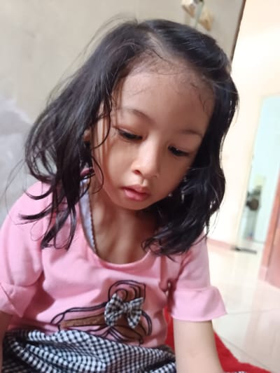 An indoor scene in Sindangasih, Indonesia, depicts a young East Asian female child, estimated to be between 3 and 6 years old. The child has dark, wavy, shoulder-length hair and is positioned with her head tilted down, her gaze directed downwards, indicating focus on an unseen element below the frame. She wears a pink short-sleeved t-shirt adorned with a brown graphic design featuring a black-and-white checkered bow. A white undergarment with a scalloped lace trim is visible beneath the neckline of the t-shirt. Her lower body is clad in a black-and-white checkered garment, consistent with a skirt or shorts. The child is situated on a red textile surface, possibly a rug or mat, partially visible at the bottom right. The background on the left consists of a light-colored, textured wall. To the right, a doorway reveals an adjacent room with light-colored, possibly tiled, flooring. A light-toned object is affixed to the upper right wall or ceiling in the background.