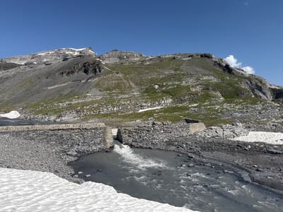 The image depicts a natural mountain landscape under a clear blue sky, likely in the alpine region of Leukerbad, Switzerland. In the foreground, a rocky river flows, its surface agitated and showing whitecaps. The riverbanks are lined with dark grey rocks and some patches of snow. A small, stone-built structure appears to be a dam or a water diversion point, with water visibly flowing through an opening.
Further back, a rough stone wall runs horizontally across the scene, separating the river area from what appears to be a road or pathway. The mountains in the background are characterized by steep, grey rocky slopes interspersed with patches of green vegetation and lingering snow. The upper reaches of the mountains are capped with snow. The overall impression is one of a rugged, high-altitude environment. No people or animals are visible in the image.