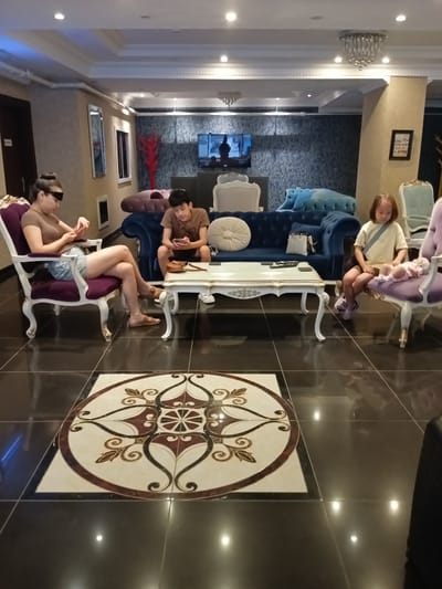 The image depicts an indoor waiting area with a modern, luxurious aesthetic. In the foreground, a woman with her hair in a bun sits on a purple upholstered armchair, wearing a brown t-shirt and denim shorts. She is looking at her phone, which she holds in her right hand. Beside her, a young boy sits on a blue sofa, also looking at a phone. In the background, a young girl sits on a purple upholstered chair, facing away from the camera, with a baby lying on her lap. The room features dark, reflective tile flooring with an ornate circular inlay. Plush sofas and ornate armchairs are arranged around a white coffee table. The walls are decorated with patterned wallpaper and framed pictures, and a television screen is visible in the background. The lighting appears to be a mix of recessed ceiling lights and decorative chandeliers. The overall impression is one of a comfortable and stylish waiting room. The specific location is an unknown facility in Kırıkkale, Turkey.