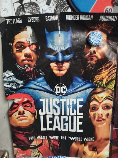 The image displays a portion of a "Justice League" movie poster, likely a promotional item. The poster features prominent images of the main characters: Cyborg on the left, Batman in the center, Aquaman on the right, Wonder Woman at the bottom right, and The Flash at the bottom left. The title "JUSTICE LEAGUE" is emblazoned across the center. Above the character images, their names are listed: "THE FLASH," "CYBORG," "BATMAN," "WONDER WOMAN," and "AQUAMAN." A tagline below the title reads "YOU CAN'T SAVE THE WORLD ALONE." The poster appears to be attached to a wall, with other papers and drawings visible in the background. The scene is indoors, with neutral lighting. No specific location in the city of Werl, Germany, can be determined from this image.
