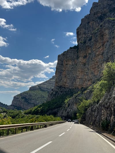 The image depicts a road in Donje Mrke, Montenegro, winding through a narrow gorge. A sheer rock face rises on the left side of the road, contrasting with a blue sky dotted with white clouds. Vegetation clings to the rock face in places. On the right side of the road, a metal guardrail separates the road from a steep drop-off covered in dense greenery. Several cars are parked on the side of the road.
