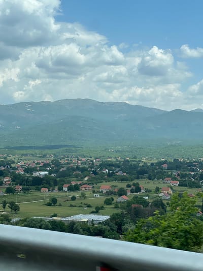 The image depicts a view in Miločani, Montenegro, overlooking a landscape of houses, trees, and mountains. A long, green mountain dominates the central part of the image. Clouds fill the sky on the left side of the image. Scattered houses with red roofs are visible among trees and greenery. A grey metal rail is partially visible in the bottom right corner.