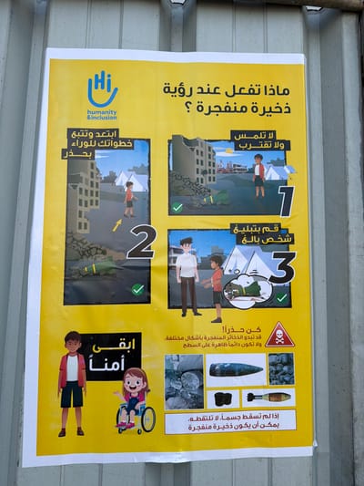 The image displays a yellow public safety poster affixed to a grey wall in a-Zawayda, Palestinian Territory. The poster, created by "hi humanity & inclusion," is an educational guide on what to do when encountering unexploded ordnance (UXO).

The central question in Arabic reads: "ماذا تفعل عند رؤية ذخيرة منفجرة ؟" (What do you do when you see unexploded ordnance?). The poster provides three illustrated steps with cartoon figures in a backdrop of damaged buildings and rubble, indicating a conflict-affected environment:
1.  **"لا تلمس ولا تقترب" (Do not touch and do not approach):** A boy stands near an unexploded bomb.
2.  **"ابتعد وتتبع خطواتك للوراء بحذر" (Step back and retrace your steps carefully):** The boy is shown walking away from the UXO.
3.  **"قم بتبليغ شخص بالغ" (Report to an adult):** The boy points out the UXO to an adult.

A warning section with a skull and crossbones symbol advises: "كن حذراً! قد تبدو الذخائر المنفجرة بأشكال مختلفة. ولا تكون دائماً ظاهرة على السطح" (Be careful! Unexploded ordnance may appear in different shapes. And it may not always be visible on the surface). This section includes photographic examples of various UXO types, such as mortar shells, artillery shells, and fragments. Another warning states: "إذا لم تسقط جسماً، لا تلتقطه. يمكن أن يكون ذخيرة منفجرة" (If it's not a dropped object, don't pick it up. It could be unexploded ordnance).

The bottom right corner features the "hi humanity & inclusion" logo and the phrase "ابقى أمناً" (Stay safe), accompanied by cartoon images of a smiling boy and a smiling girl in a wheelchair. The poster's clear graphics and concise Arabic text aim to provide vital safety instructions to the public in a post-conflict or hazard zone.