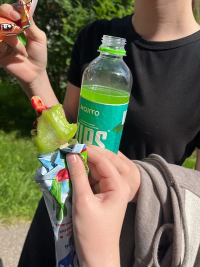 Two individuals are outdoors, engaged in consumption of frozen treats and beverages. In the foreground, a person's hands are holding a melting green popsicle with a red center, presented in a crumpled, colorful wrapper adorned with images of cacti and cherries. To the right, another person's hands hold a transparent plastic bottle containing a vibrant green liquid, labeled "MOJITO" in Russian. The background features blurred green foliage and sunlight, suggesting a park or a similar outdoor setting. The overall scene depicts a casual, warm-weather moment.