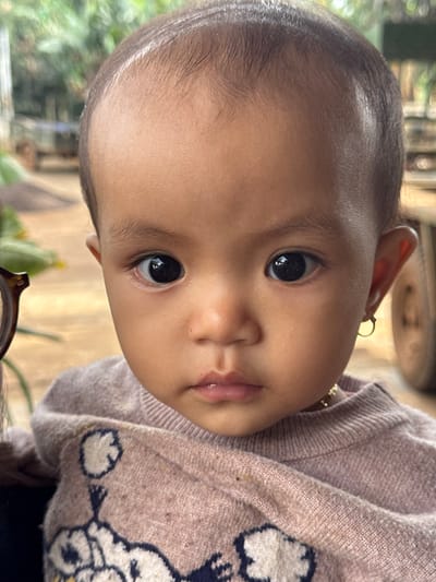 The image displays a close-up portrait of a young child, likely an infant or toddler, with a medium complexion, dark hair, and large, dark eyes, looking directly at the viewer. The child is wearing a light pink or lavender long-sleeved sweater featuring a white and dark patterned design. A small gold-colored hoop earring is present in the child's right earlobe, and a thin gold-colored chain is visible around the neck. On the far left, a fragment of brown-rimmed eyeglasses is visible, suggesting the presence of an individual holding the child. The background is out of focus, depicting an outdoor environment in Ko H'Neh, Vietnam, with green foliage on the left and blurred structures or objects, including what appears to be a vehicle tire or part of a building, on the right. The distant ground appears unpaved.
