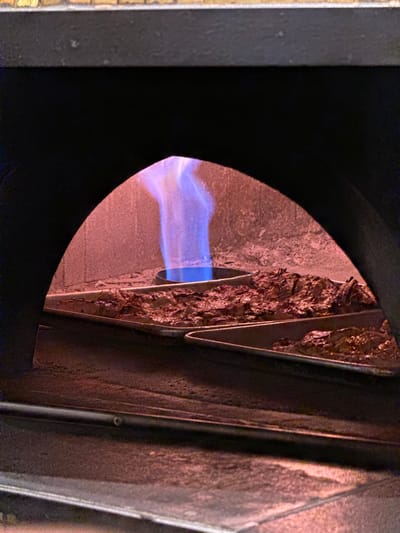 A blue flame is visible inside an oven, with trays of meat cooking on racks below. The oven appears to be made of brick and metal. The scene is lit with a reddish glow from the heat of the oven. This image was taken in West Palm Beach, United States of America.