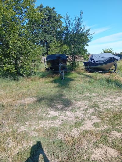 The image depicts an outdoor scene in Sokyrna, Ukraine, with two boats on trailers parked in a grassy area surrounded by trees under a clear blue sky. The boat on the left is dark and uncovered, with a trailer visible. The boat on the right is covered with a dark grey tarp. The ground is covered with dry grass and patches of bare soil. The foreground shows the shadow of a person taking the photograph. The environment suggests a storage or recreational area, possibly near a body of water, given the presence of boats.