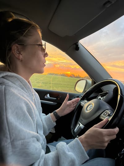 A medium shot captures a woman driving a car at sunset. She is wearing a grey hooded sweatshirt and glasses, and her hair is pulled back. Her hands are on the steering wheel, and she is looking out the windshield at the vibrant orange and yellow sky, which is dotted with clouds. The car's interior is visible, including the dashboard and the driver's side door. Outside the car, a green field with some trees is visible in the distance. The scene appears to be set in a rural area, possibly near Perevoznoye, Russia, given the time of day and the landscape.