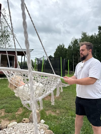A man in a white t-shirt and black shorts stands in a grassy yard, holding the ropes of a white macrame hammock. A child with blonde curly hair lies in the hammock, dressed in white. In the background, a small red building with a porch is visible, along with a green trampoline and trees under a cloudy sky. The location is the city of Ryzhkovo, Russia.