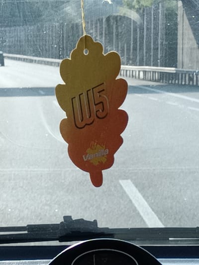 The media file shows a car air freshener hanging from a string inside a vehicle. The air freshener is leaf-shaped and features the text "W5" prominently in large, stylized letters. Below "W5", the word "Vanilla" is visible in smaller lettering, along with a graphic representation of a leaf. The air freshener transitions in color from yellow at the top to orange at the bottom. The background, seen through the car's windshield, depicts a road with white lane markings and a guardrail. Trees and a concrete structure are also visible in the distance. The lighting suggests it is daytime. The overall scene is typical of being in a car traveling on a road.