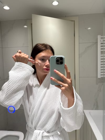 A young female, wearing a white waffle-knit bathrobe and a gold-colored ring on her left ring finger, takes a mirror selfie in a modern bathroom. She holds a light blue iPhone in her right hand, reflecting its triple-lens camera array. With her left hand, she holds a rose gold-colored cosmetic applicator, applying a small dot of white cream or serum to her left cheek. The bathroom features white tiled walls with a grey marble-like pattern, a white ceiling with recessed circular lights, a partially open plain white door reflected behind her, a white toilet in the bottom left foreground, a white bathtub on the right, and a white rectangular towel warmer mounted on the right wall.