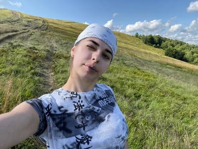A young woman, wearing a light blue bandana with a white paisley pattern and a grey t-shirt with a black graphic print, takes a selfie on a grassy hillside. The woman is looking towards the camera, with her left eye partially closed. Her lips are parted, and she appears to be mid-speech or in the process of blinking. The background consists of a rolling green hillside with a dirt track winding up it, and a distant treeline on the right side of the frame. The sky above is blue with scattered white clouds. The image appears to have been taken outdoors, in a natural, possibly mountainous, environment, likely in the vicinity of Ust'-Chorna, Ukraine, given the landscape features.