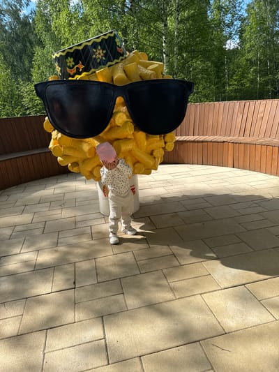 A child in a pink cap and white clothing is standing next to a large sculpture of french fries with sunglasses in Zelenodolsk, Russia. The sculpture is painted yellow and is next to a wooden wall. The ground is paved with rectangular stones. Trees are visible in the background.