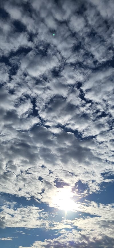 This image captures a dynamic sky filled with a mixture of fluffy white clouds and darker, heavier cloud formations against a vibrant blue sky. The sun is visible in the upper right quadrant of the image, shining brightly and creating a lens flare effect. The clouds appear to be moving or forming, suggesting a day with changing weather conditions. The orientation of the image is vertical, with the sun appearing on the right side. There is a small, circular, bright turquoise object visible in the sky, positioned to the left of the center. This could be a photographic artifact or a distant object. Another small dark object, possibly a bird or insect, is visible in the upper left portion of the frame. The scene evokes a sense of openness and the vastness of the sky. The lighting suggests it is likely daytime, with the sun's position indicating it could be morning or afternoon. No text is visible in the image. The location context provided is Hetmanivka, Ukraine.