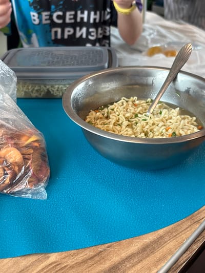 The scene, located in Ivanovka, Russia, depicts an indoor setting focused on a table surface. A stainless steel bowl, exhibiting condensation on its exterior, is positioned prominently on a bright blue, textured placemat. Inside the bowl, prepared instant noodles with visible green and orange dehydrated vegetable fragments are immersed in broth, and a silver spoon with a fluted handle rests within. To the left of the bowl, a transparent plastic bag contains numerous dried, reddish-brown, and yellow fruit or vegetable pieces. Behind these items, a grey rectangular plastic container with a patterned, semi-transparent lid is also visible, containing what appears to be instant noodles. The blue placemat rests on a wooden-patterned table. In the upper background, the upper torso and arms of an individual are partially visible. The person is wearing a dark blue T-shirt featuring prominent white Cyrillic text that reads "ВЕСЕННИЙ ПРИЗЫВ" (VESENNIY PRIZYV), translating to "Spring Conscription" or "Spring Draft." A yellow wristband is present on the individual's right wrist. The background beyond the person is indistinct, appearing as a blurred white fabric.