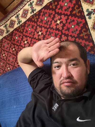 In Yabogan, Russia, a person is lying down on a blue, ribbed surface, possibly a sofa. They are wearing a black t-shirt with a white Nike logo on the left chest and "NIKE" written on a patch near the collar. The person has their right arm raised, with their hand resting on their forehead. They have short, dark hair and stubble on their face. Behind them, a richly patterned rug with red, beige, and green geometric designs is visible. The lighting suggests an indoor setting.