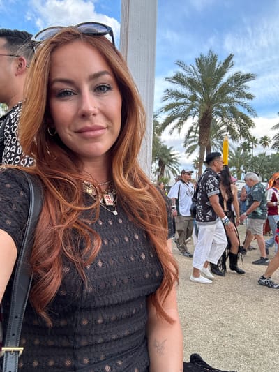 The image shows a selfie of a woman with long auburn hair, wearing a black lace-patterned top and multiple necklaces. Sunglasses are perched on her head. A tattoo is visible on her upper arm. In the background, blurred figures of people walking are visible, some wearing casual clothing like shorts, hats, and patterned shirts. Palm trees are present, and a bright blue sky is seen with some white clouds. Given the palm trees and the presence of crowds in casual clothing, it is likely that this photo was taken in Coachella, California, United States of America.