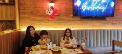 A family celebrates a birthday in a restaurant. A woman sits beside a boy eating pizza. Next to him, a girl with heart-shaped sunglasses holds a slice of pizza. On the table is an assortment of pasta and salad dishes. Red and white balloons are above them. A TV displaying "Happy Birthday!" is mounted on a wooden wall.