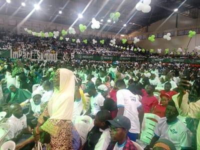 This image depicts a densely populated indoor event held in a large hall or arena in Ushapa, Nigeria. The venue features a high ceiling with visible industrial lighting fixtures and tiered seating in the background, which is largely occupied by attendees. The foreground shows a crowded floor level where individuals are seated on white plastic chairs.
A prominent banner displays "Impact Summit 2015" and multiple "NEOLIFE" logos (a green circle with stylized leaves). Green and white balloons, often grouped in threes, are suspended from the ceiling and adorn the upper railings. Many attendees are dressed in white or light-colored shirts, some bearing logos consistent with the banners. Numerous women are present, many wearing head coverings, including green and white hijabs/scarves.
In the mid-foreground, a woman in a patterned brown, black, and white dress with gold accents, wearing a light yellow headscarf, stands or leans slightly, looking to her left. To her lower left, a bald man with a beard, wearing a white polo shirt, is seated, holding a white object. Other individuals in the foreground include a young man in a black baseball cap and plaid shirt, and several people in white t-shirts with blue or green accents, seated and observing the event. The crowd is oriented towards an unseen focal point, suggesting an ongoing presentation or performance.
