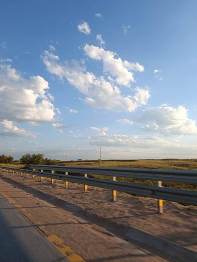 The image captures a roadside scene under a partly cloudy sky. In the foreground, a yellow and grey striped road divider runs along the edge of a paved road. Beyond the divider, a vast, dry field stretches towards the horizon, dotted with scattered trees on the left and utility poles in the distance. The sky is a vibrant blue, filled with scattered white cumulus clouds. The lighting suggests it is late afternoon or early evening, casting long shadows. The location is identified as the city of Rassvet, Kazakhstan. There are no people or animals visible in the image.