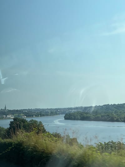 The image shows a wide river, likely the River Foyle, in the city of Derry/Londonderry, United Kingdom. On the left bank of the river, there is a view of a cityscape with several buildings, including a prominent church spire. The cityscape is nestled amongst lush green trees. The river itself is calm and reflects the light blue sky. On the right bank of the river, there is a dense area of trees. The foreground of the image is blurred, suggesting it was taken from a moving vehicle, possibly a train or car. The sky is mostly clear with some faint wispy clouds and some reflections on the window. The overall impression is a scenic view of a river passing through a city surrounded by nature.