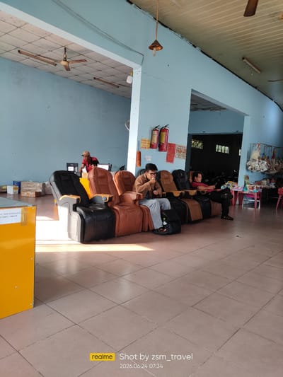 This image captures a waiting area, possibly at a bus station or transportation hub, in Gò Dầu, Vietnam. The scene is dominated by several large, reclining massage chairs, predominantly in brown and black leather. Five individuals are visible; four are seated in the chairs, with one person standing in the background near a counter.
One man, wearing a red shirt and a beige baseball cap, stands behind a yellow counter, appearing to interact with a computer monitor. Next to him, a young man in a brown jacket and jeans is seated in a chair, looking down. To his right, another man in a red shirt and black pants is seated, looking at his phone. Further down the row, a man in a brown jacket and light blue jeans is seated, holding a piece of food. On the far right, a woman in a black top and dark pants is seated in a chair, looking towards the right.
The environment features pale blue walls and a tiled floor. Several objects are present, including fire extinguishers mounted on the wall, boxes stacked in the background, and a display of items hanging on a rack on the right side of the frame. A ceiling fan is visible, and sunlight streams into the room, casting shadows on the floor. A yellow structure, possibly a service counter or kiosk, is prominently positioned on the left. The overall impression is one of a functional, albeit somewhat dated, waiting area.