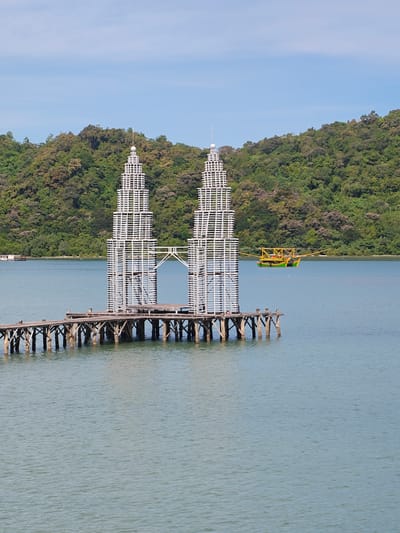 The image shows a replica of the Petronas Twin Towers made of white wooden planks, standing on a wooden pier in the water. The replica is located in Layeun, Indonesia. A boat with a yellow and green body is visible on the water behind the replica. A wooded shoreline is in the background. The sky is blue.