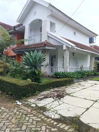 The scene captures a two-story residential building, purportedly located in Pô Kô, Vietnam, under an overcast sky. The primary structure features a white facade and a reddish-brown tiled roof. The second story includes an arched balcony with a silver-colored metal railing. The ground floor presents a covered porch supported by white columns and enclosed by white balustrades, with a dark wall-mounted lantern visible.

In the foreground, a concrete paved area exhibits extensive cracking and is partially overgrown with grass and weeds. Sections of the paving consist of square slabs, while an area to the left features interlocking pavers. Dry plant debris is scattered across the cracked pavement, and a small piece of white and red litter is visible on the interlocking pavers.

Adjacent to the paved area, a manicured green hedge forms a boundary for a garden. Within the garden, a large, prominent agave-like succulent plant with broad green leaves stands centrally. Smaller agave-like plants line the perimeter of the front porch. Various other green foliage is also present in the garden space.

To the left of the main white building, a portion of an orange-colored house with a tiled roof is visible. To the right, in the background, a blue water tank mounted on a metal stand is discernible. No individuals are present in the image.