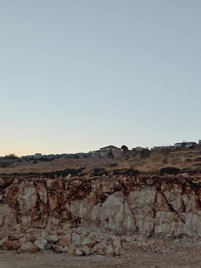 The image depicts a rocky, excavated area in the foreground, with layers of light-colored stone and darker soil. Red chalk markings, including numbers and a character that appears to be a Chinese character, are visible on the rock face. Beyond the excavation, a hillside is covered with dry vegetation and scattered debris, leading up to a cluster of residential buildings. The buildings are varied in style and color, with some appearing to be single-family homes and others possibly multi-unit dwellings. Power lines and utility poles are also present, crisscrossing the landscape. In the distance, the sky is a pale blue, suggesting either early morning or late evening. The scene appears to be a construction or excavation site located on the outskirts of a settlement or urban area. Given the context provided, this scene is situated within the Qalandiya Refugee Camp, Palestinian Territories.