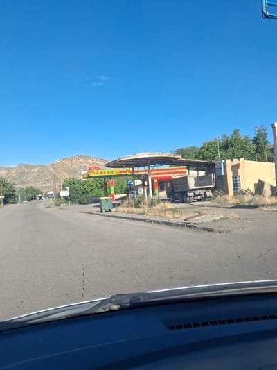 A wide-angle, eye-level shot captures a rural street scene in Vayk, Armenia, under a clear blue sky. On the right, a gas station with a covered canopy is visible. The canopy's roof is supported by red and green striped pillars and is covered with what appears to be a weathered, textured material. The gas station building is a single-story structure with tan walls, featuring a few windows. To the left of the gas station, a green dumpster sits near the edge of the road. A large, grey dump truck is parked under the canopy of the gas station. The road surface is asphalt, with some cracks and debris. In the background, a dry, rocky hillside rises, dotted with sparse vegetation. Trees line the left side of the road, and further in the distance, other structures and power lines are visible. The foreground of the image shows the dashboard and windshield of a vehicle.
