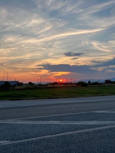 The image captures a sunset over a rural landscape in Chaidefto, Greece. The sky is filled with wispy clouds painted in hues of orange, yellow, and blue, indicating the setting sun. The sun itself is a small, bright red orb visible on the horizon.

In the middle ground, a grassy field stretches out, bordered by a line of trees and utility poles. Beyond the field, some buildings and distant mountains can be seen. One building in the distance has text on it that appears to read "MARINE CENTER".

To the right of the field is a paved road, possibly a highway or a long, straight road, with distinct white lines marking the lanes. The road surface is dark asphalt with visible aggregate.

The overall scene evokes a sense of tranquility and the beauty of nature at dusk. The weather appears clear, with the dramatic cloud formations suggesting a beautiful end to the day. The time of day is clearly sunset, given the color of the sky and the position of the sun. There are no people visible in the image.