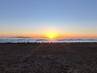 An unobstructed wide shot captures a serene sunset scene at the beach in Agios Stefanos, Greece. The sky is a gradient of soft blues and oranges, transitioning to a brilliant yellow at the horizon where the sun is a bright disc. The sun casts a shimmering reflection on the calm sea. A distant island or landmass is visible on the horizon. In the foreground, a dark, sandy beach stretches out, with a line of dark rocks separating it from the water. A few small birds are silhouetted against the sky, indicating their flight. The overall atmosphere is tranquil and natural.