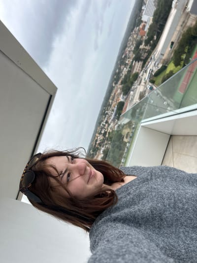 The image captures a young female positioned on an elevated balcony in Les Meravelles, Spain, against a panoramic cityscape. The female, in the foreground, has shoulder-length dark brown hair and is wearing a grey textured sweater. Tortoiseshell-patterned sunglasses are resting on top of her head. Her face is directed towards the camera, exhibiting a slight smile. Behind her, a clear glass railing with a white frame borders the balcony, offering a view of a dense urban environment under an overcast sky. The cityscape includes numerous buildings of varying sizes, roads, and interspersed green areas. Specifically, a sports complex with at least two red-surfaced tennis courts and a green field is visible in the mid-ground. The balcony floor is a light-colored, possibly tiled, surface.