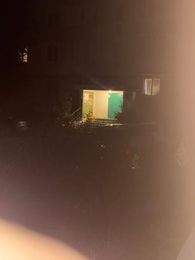 The image depicts a nighttime scene in the city of Skalyste, Ukraine. In the center of the frame, an entrance to a building is visible, illuminated by an overhead light and light emanating from within. The entrance features a green door and a set of stairs leading up to it. There are posters or notices displayed on the wall next to the door. To the right of the entrance, a window with two panes of glass is illuminated. To the left of the entrance, a small, dark-colored car is parked. The foreground and surrounding areas are obscured by darkness and foliage, with some faint lights suggesting other structures or activity in the distance. The overall impression is one of a residential or communal building entrance at night.