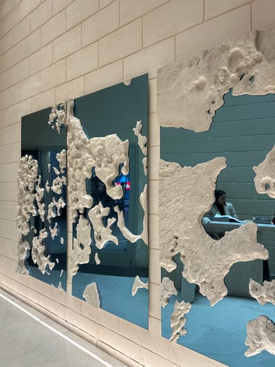 Three rectangular, blue-tinted mirrors are arranged sequentially on a ceiling with textured, white borders resembling clouds. The ceiling itself is composed of beige bricks laid in a horizontal pattern. Reflections within the mirrors show parts of the surrounding environment. A woman is visible in the reflection of the top mirror. The reflective surfaces and cloudy borders create a surreal, artistic ceiling installation.