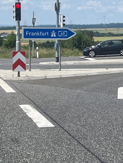 The image shows a street sign indicating "Frankfurt" and the highway number "49" in Stadtallendorf, Germany. The sign is blue and white with an icon of a bridge and a car. Next to it is a red traffic light, indicating a stop. A red and white chevron sign points to the left, suggesting a turn or curve in the road. In the background, a dark-colored car is partially visible on the road. Beyond the road, a landscape of fields, trees, and wind turbines can be seen under a partly cloudy sky. The lighting suggests it is daytime, likely late morning or afternoon, given the shadows cast on the road. The scene appears to be a highway interchange or a point where a road transitions into a highway. There is no visible text beyond the signage.