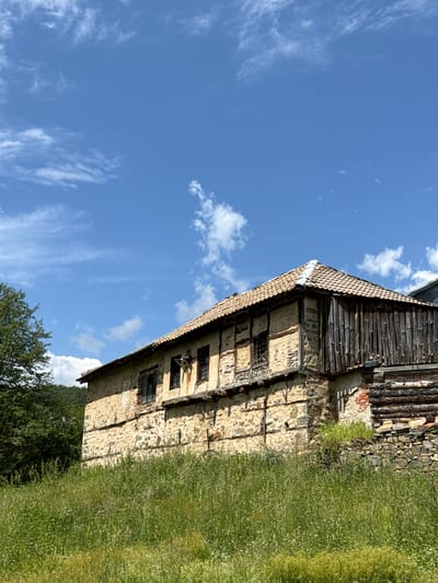 The image shows an old, stone building with a wooden upper section in Ogradna, Bulgaria. The building has a tiled roof and several windows, some of which are boarded up or broken. The building is bordered by green grass and trees. The sky is blue with wispy clouds.