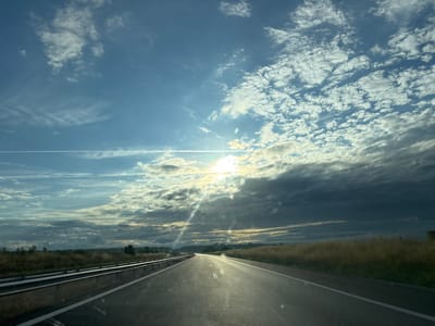 The image captures a highway driving perspective with a dramatic sky. The road stretches forward, seemingly curving slightly to the right, marked with white dashed lines. On the left, a metal guardrail runs parallel to the road, with a concrete barrier beneath it. The roadside is lined with dry, golden grass and sparse trees in the distance. The sky is filled with a dynamic mix of clouds: bright white, scattered cumulus clouds higher up, and darker, more ominous clouds lower towards the horizon. Sunlight breaks through in the upper right portion of the sky, creating a radiant glow and casting light rays that are visible on the road surface and through the windshield. A faint contrail from an aircraft is visible across the upper left sky. The scene appears to be set during the daytime, with the sun positioned at an angle that suggests either early morning or late afternoon. There are no people or vehicles visible in the immediate frame, emphasizing the sense of travel and the vastness of the open road. The location is identified as Kolarovo, Bulgaria.