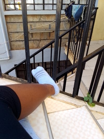 A dark-skinned leg, clad in black fabric (shorts or trousers) and a light blue sock with three horizontal grey stripes, is positioned on a light-colored, square-tiled surface featuring a subtle checkered pattern and visible grout lines. A small green plant grows from the tiled surface near the base of a black metal staircase railing, which extends downwards and to the right, indicating a multi-level structure.

The background reveals a wall constructed from light brown/beige concrete blocks with horizontal indentations. Dark vertical stains or drips are visible below a mounted metallic bracket/shelf on this wall. Above the concrete blocks, a window secured with a metal grille is present. A white vertical pipe runs along the left side of the scene. Several pieces of blue and dark fabric items are draped over the upper section of the railing to the right. Thin wires or strings are also discernible in the background, adjacent to a light yellow wall section on the far right. The scene depicts an outdoor or semi-outdoor area, likely a building's landing, located in Kalimoni ward, Kenya.