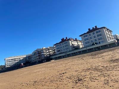 The image depicts a sunny day on Zarautz Beach in Zarautz, Spain, viewed from the sandy shore towards the urbanized coastline. The foreground is dominated by extensive light-brown sand, marked with numerous footprints. In the mid-ground, a single individual, dressed in dark attire, stands on the beach facing right, near the retaining wall that separates the beach from an elevated promenade. Along the promenade, multiple smaller, indistinct figures are visible. A row of multi-story residential or hotel buildings with varying architectural styles lines the coast in the background. Several buildings feature light-colored facades and prominent red-tiled roofs, while others present more modern designs with balconies. A red and yellow playground structure is visible on the embankment in front of one of the buildings on the left. The sky is a clear, uninterrupted bright blue, occupying the upper half of the frame. The entire scene is brightly lit by direct sunlight.