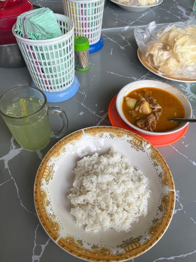 An overhead shot captures a prepared meal set on a dark grey table with white marble-like veining. In the foreground, a large, ornate white plate with a gold and brown patterned rim holds a mound of fluffy white rice. To its left, a white bowl filled with a rich orange-brown stew containing chunks of meat and possibly potatoes is placed on a bright orange saucer, with a spoon resting in the bowl.
Above the stew bowl, another white bowl, partially visible, contains a pile of light-colored, crispy crackers, some of which are still enclosed in a clear plastic bag. To the lower left, two cylindrical white plastic mesh holders are seen, one containing neatly folded green napkins and the other with similar pink napkins. One of these holders has a blue base, and the other has a red base. A small, clear container with a green lid, filled with toothpicks, is placed between these holders.
Further to the right, a clear glass mug with a handle contains a light green beverage with ice and possibly some seeds, and a yellow straw is visible inside. The overall scene suggests a casual meal, likely served for multiple people, given the multiple napkin holders and the serving sizes. There is no visible text.