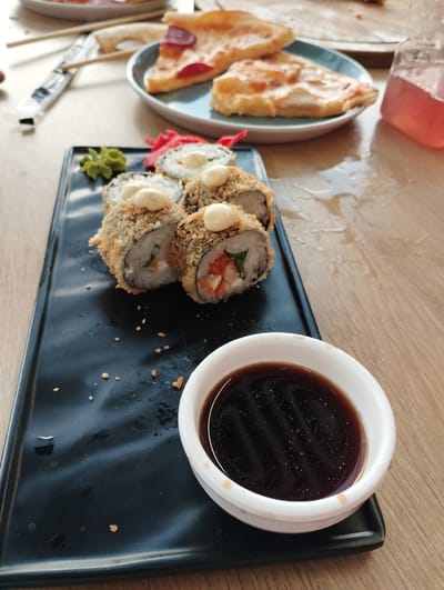 In a casual dining setting, possibly a cafe or restaurant in Talas City, Kyrgyzstan, a close-up shot focuses on a meal. A black rectangular plate holds several pieces of fried sushi rolls, topped with a white sauce and a crispy coating. Next to the sushi is a small mound of green wasabi and some bright pink pickled ginger. To the right, a small white dipping bowl contains dark soy sauce. In the background, out of focus, are slices of pizza on a blue plate and a glass of pink beverage. The table surface is a light-colored wood. The overall impression is of a shared meal, with a focus on the prepared food.