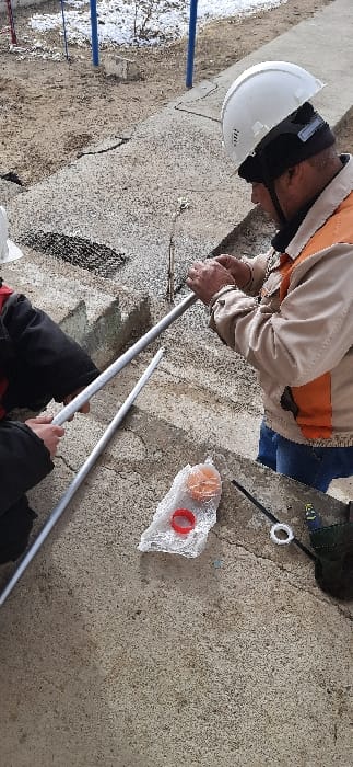 A worker wearing a hard hat, safety glasses, and an orange vest is seen connecting two silver pipes together. Another person, only visible from the waist down, is holding one of the pipes. An orange and white object is wrapped in clear plastic near a black tool and a white ring. The ground is concrete. In the background, there is a small blue fence and some dirt and snow.