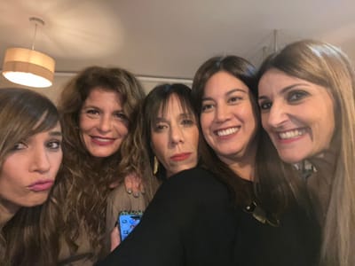A close-up, indoor group selfie depicts five adult women, estimated to be in their 30s to 50s, positioned in a tight formation, looking towards the camera. From left to right: a woman with long brown hair and bangs pouts; a woman with curly brown hair smiles broadly, her left arm extended to the first woman's shoulder, holding a smartphone with visible app icons, likely taking the photo; behind them, a woman with dark hair and bangs shows a neutral expression; next, a woman with dark brown hair smiles broadly, revealing teeth, wearing a dark top and a dark pendant necklace; and finally, a woman with long light brown hair smiles broadly, revealing teeth. The women generally wear red lipstick. The background is a blurred interior space with a light-colored ceiling, from which a light-colored cylindrical ceiling fixture hangs in the upper left. The lighting is warm. The exact location within Alcalá De Henares, Spain, is not discernible from the image.