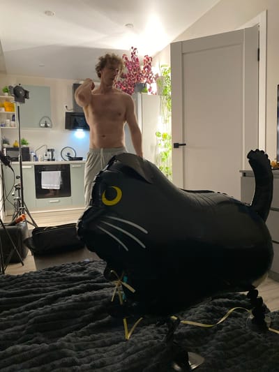 The image depicts an indoor scene, possibly a kitchen or living area. In the foreground, a large black inflatable cat balloon is prominently displayed, resting on a textured gray rug. The cat balloon has yellow eyes and white whiskers. Behind the balloon, a shirtless man with curly blond hair is standing and gesturing with his right arm. He is wearing light gray sweatpants. The background reveals a kitchen area with cabinets, appliances, and shelves stocked with various items. A camera light stand is positioned to the left of the man. To the right, a white door is visible, and further back, several plants are arranged on shelves. The overall lighting suggests an indoor setting at dusk or night. There is no indication of the city of Rahya, Russia, in the image.