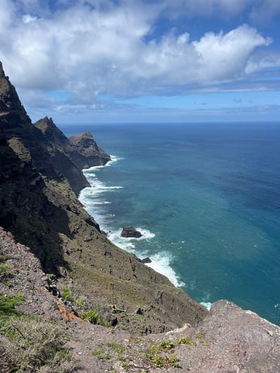 A high-angle shot depicts a rugged coastline in Agaete, Spain, with steep cliffs plunging into the Atlantic Ocean. The cliffs are primarily rocky and covered with sparse vegetation. Waves crash against the rocks below, creating white foam. In the distance, the ocean transitions from deep blue to a lighter shade near the horizon, which blends with the sky. The sky is partly cloudy with patches of blue. No people are visible in the image.
