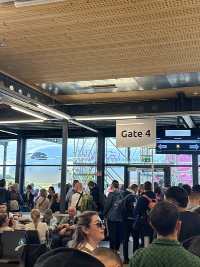 The image depicts a busy airport gate area with a large crowd of people waiting. A sign reads "Gate 4" and an electronic display shows flight information for Wizz Air to Sofia (SOF), with "Boarding" indicated. An airplane is visible through the large windows, and boarding stairs are set up. The environment appears to be an indoor airport terminal with modern architecture, including a perforated ceiling and metal support beams. People are dressed casually, many are carrying luggage or backpacks, and some are seated. The overall atmosphere is one of transit and anticipation for travel. The location of Künersberg, Germany is not discernible from the visual information in the image.